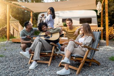 Gitar çalan, şarkı söyleyen, evcil köpeği tutan, karavanın yanındaki şezlonglarda oturan, birlikte kamp yapan, sonbahar tatilinde eğlenen, kırsalda vakit geçiren çok ırklı genç arkadaşlar.