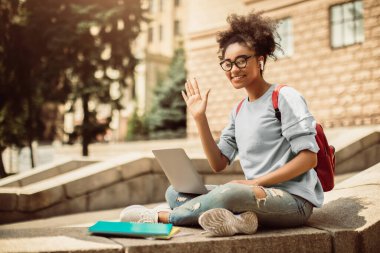E-öğrenme. Bilgisayarlı, el sallayan, kameraya gülümseyen, kolej binasının önünde oturan siyah öğrenci kız. Uzaklık Eğitimi ve Modern İletişim Konsepti