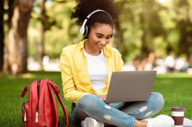 Neşeli Afrikalı Öğrenci Kız Laptop 'ta öğreniyor. Dışarıda park yerinde oturan kablosuz kulaklıkla video dersi izliyor. Modern Eğitim, Uzaklık Çalışması ve E-Öğrenme Konsepti