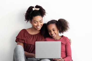 Mutlu siyah anne ve genç kız dizüstü bilgisayar kullanıyor, beyaz duvarın üzerinde oturuyor, Afrikalı Amerikalı kadın ve kız ellerinde defter tutuyorlar, internetten film izliyorlar, reklam için yer kopyalıyorlar.