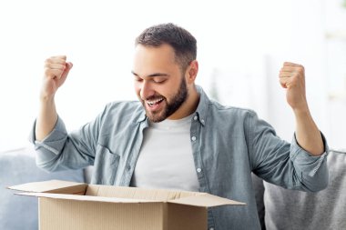 Tatmin olmuş erkek müşteri karton kutuyu açıyor, teslimat için heyecanlanıyor, çevrimiçi mağaza siparişi alıyor, evde EVET hareketi yapıyor. Oturma odasında paketleri açan genç adam.