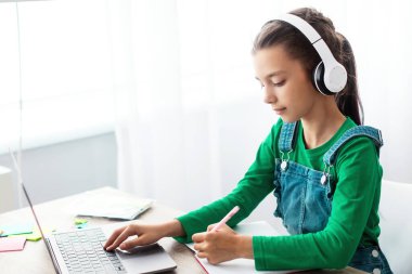 Eğitim Dersi Konsepti. Kablosuz kulaklıklı çalışkan genç bir kızın portresi. Laptop kullanıyor, deftere yazı yazıyor, öğretmenini dinliyor, bedava fotokopi çekiyor.