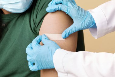 İş kıyafetleri hemşiresi, yüz maskesi takan genç Hintli adam için koronavirüs aşısı yapan eldivenler, omuza yapıştırıcı bant takmak, bej rengi stüdyo arka planı, yakın plan. Güvenlik aşısı kavramı