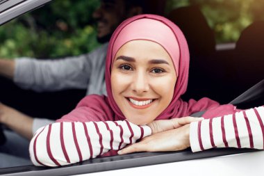 Arabasının camından sarkan ve kameraya bakan, orta doğulu gülümseyen, yolcu koltuğunda oturan, kocasıyla otomobil yolculuğunun keyfini çıkaran tesettürlü güzel müslüman kadın portresi.
