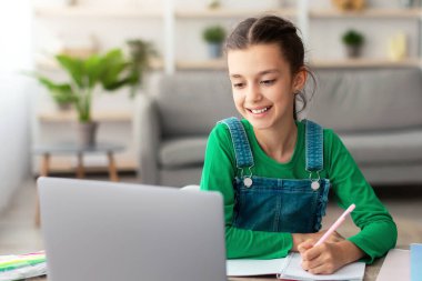 Online Eğitim Konsepti. Masasında oturan gülümseyen genç kız dizüstü bilgisayarına bakarak not defterinde yazıyor. Öğrenci kız çevrimiçi kurs izliyor, not alıyor, ödev yapıyor.