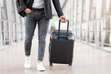 El çantalı tanınmayan bir adam havaalanı terminalinde yürürken günlük kıyafetleriyle uçağa bagajla binen, iş seyahatine çıkmaya hazır genç bir adamın kırpılmış fotoğrafı.