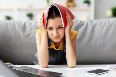 Okul stresi konsepti. Üzgün öğrenci kız defterini kafasında tutarak masadaki koltukta oturuyor, ödevlerini yapıyor, görev yüzünden bunalımda, oturma odasında uzaktan ders çalışan genç kız. Ev Tabanlı Öğrenme