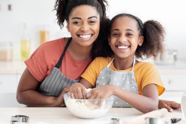 Neşeli Afro-Amerikan ailesi genç anne ve genç kız evde kurabiye yapıyorlar, önlük giyiyorlar, ev yapımı bisküviler veya turtalar için hamurla çalışıyorlar, kameraya sarılıyorlar ve gülümsüyorlar.