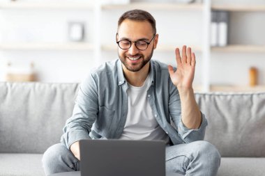 Çevrimiçi toplantı. Laptop kullanan mutlu beyaz bir adam, video görüşmesi ya da iş görüşmesi yapıyor, içeride web kamerasına el sallıyor. Millennial elemanı iş arkadaşlarıyla uzaktan iletişim kuruyor.
