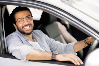 Araba kiralama, satın alma, perakende, kiralama konsepti. Mutlu Arap adam portresi, günlük giysiler içinde yepyeni bir arabada oturuyor, elini direksiyona koyuyor, kameraya gülümsüyor, yan görüş, kopyalama alanı.