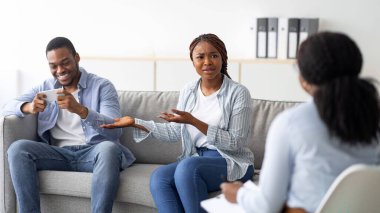 Irritated black woman complaining about her husbands smartphone addiction during psychological consultation. Professional psychotherapist trying to help young couple with gadget overuse problem clipart