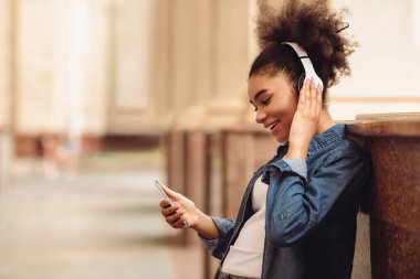 Müzik ya da Eğitim Podcast 'i dinleyen akıllı telefon kullanan siyahi öğrenci kolej binasının yanında duruyor. İnsanlar ve Gadget 'lar, Yan Görünüm