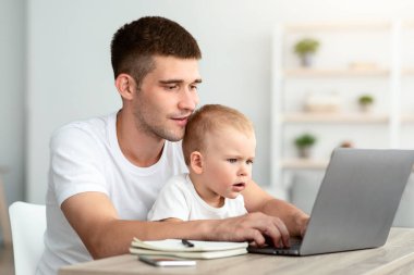 Serbest ve Çocuk Bakımı. Evde dizüstü bilgisayarda çalışan küçük bebeği olan baba, bebeğine bakıcılık yaparken bilgisayarını uzaktan kontrol etmek için kullanan genç adam, boş alanla yakın plan fotoğrafı.