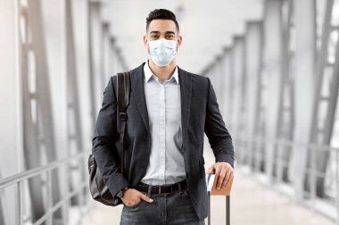 Coronovirus Seyahatleri. Tıbbi maskeli genç bir adam havaalanı binasında bavuluyla duruyor. Salgın sırasında seyahat eden orta doğulu bir adam. Pasaportu elinde biletlerle kameraya bakıyor.