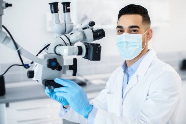 Klinikte diş mikroskobu kullanan koruyucu tıbbi maske takan Arap dişçi, hastaları tedavi etmek için modern ekipmanlarla çalışan ortadoğu stomatolog doktor, yakın çekim.