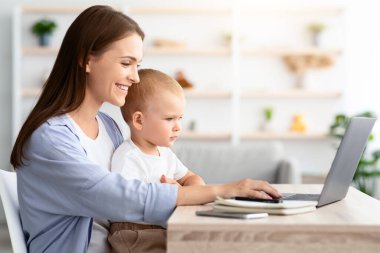 Serbest ve Doğumevi. Evdeki dizüstü bilgisayarda uzaktan çalışan ve bebeğe bakan mutlu genç bir bayan, Milenyum annesi, küçük oğluyla masada oturuyor ve bilgisayar kullanıyor, yan görüş kullanıyor, boşluğu kopyalıyor.