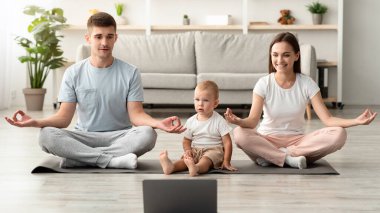 Çevrimiçi yoga. Evde dizüstü bilgisayarla birlikte meditasyon yapan mutlu ve genç bir aile, Lotus pozisyonunda oturan neşeli ebeveynler, bilgisayardaki video derslerini izliyorlar, ev içi eğitimin tadını çıkarıyorlar.