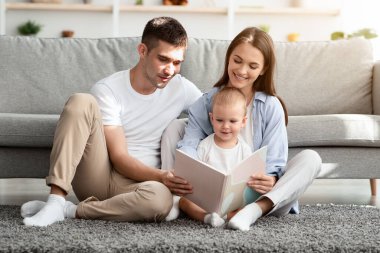 Çocuklar için Geliştirme Eğlencesi. Küçük oğluyla kitap okuyan mutlu ebeveynler, üç çocuklu Millennial aile, oturma odasında yerde dinleniyor, ev işlerinin keyfini çıkarıyor, boşluğu kopyalıyor.