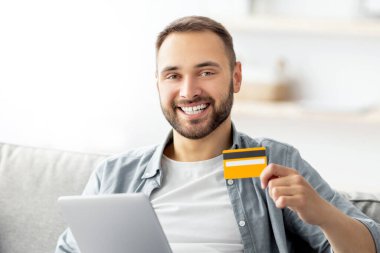 Kredi kartı ve tablet bilgisayarı olan mutlu genç adam evdeki kanepede oturup internetten mal alıyor. Yakışıklı milenyum adamı Coronavirus tecridi sırasında online mağazadan alışveriş yapıyor.