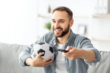Televizyonda futbol maçı izleyen, elinde uzaktan kumandayla evde top tutan genç adam. Spor fanatiği kanepede oturuyor, televizyon maçı seyrediyor, en sevdiği futbol takımı için tezahürat yapıyor.