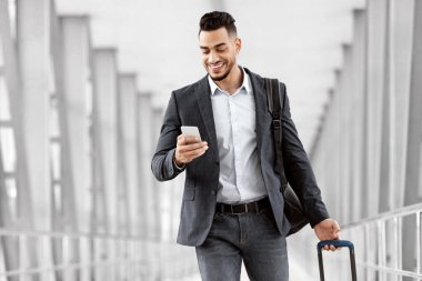Çevrimiçi Kontrol. Havalimanında çantasıyla yürüyen ve akıllı telefon kullanan yakışıklı bir Arap uçuş kapısına giderken cep telefonuyla internete göz gezdiren genç bir Orta Doğulu.