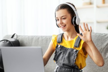 Çevrimiçi Okul Konsepti. Kablosuz kulaklık takan gülümseyen öğrenci portresi evdeki koltukta oturup ders çalışıyor bilgisayarla öğretmenle sanal görüşme yapıyor, web kameralarına el sallıyor.