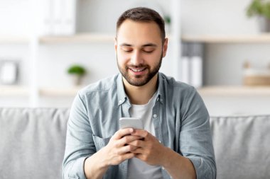 Akıllı telefonuyla kanepede oturan ve arkadaşına mesaj atan, kapalı mekanda iletişim kuran pozitif bir genç. Milenyum Kafkasyalı adam mesaj yazıyor, video görüşmesi yapıyor ya da evde sörf yapıyor.