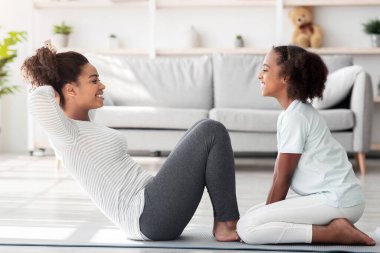 Spor kıyafetlerinde birlikte antrenman yapan neşeli siyahi anne ve çocuk spor ayakkabıları, spor paspası, karın kasları üzerinde çalışmak, mekik, yan görüş. Sportif aile annesi ve kızı spor yapıyor.