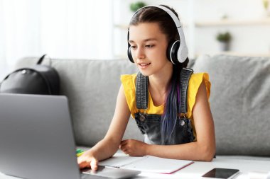 Uzaklık Eğitim Konsepti. Kulaklıklı kız çocuğunun portresi evdeki bilgisayarda çalışıyor. Boyalı saçı olan gençler ödev yapmak için dizüstü bilgisayar kullanıyor, klavyede daktilo kullanıyor.