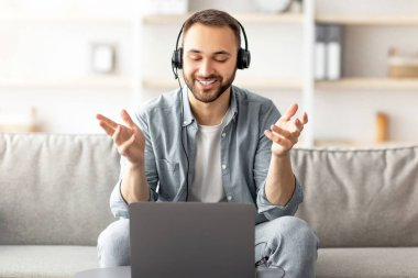 Kulaklıklı genç adam evde bilgisayarla video görüşmesi yapıyor ve kamerayla konuşuyor. Genç beyaz adam internet iletişiminden zevk alıyor, web konferansına veya iş toplantısına katılıyor