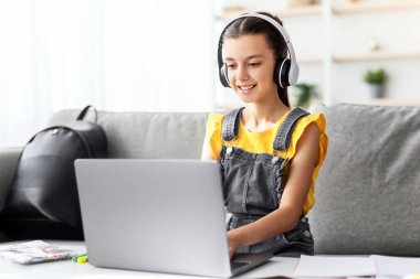 E-öğrenme konsepti. Evde dizüstü bilgisayarda çalışan kulaklık takmış gülümseyen bir genç. Mutlu gençlik ev ödevi yapmak için bilgisayar kullanıyor, klavyeye yazıyor, arkadaşlarıyla sohbet ediyor ya da ev ödevi yapıyor.