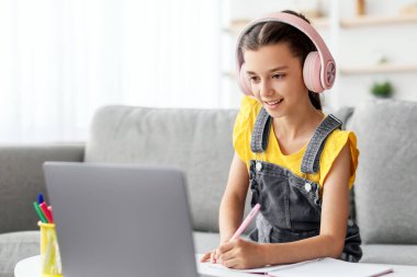 Evde ders çalışıyorum. Pembe kulaklıklı gülümseyen öğrenci masadaki koltukta oturuyor, bilgisayar kullanıyor, deneme veya sınav yazıyor, ders kitabında notlar alıyor, online ders sırasında öğretmenlerin ekranlara bakmasını dinliyor.
