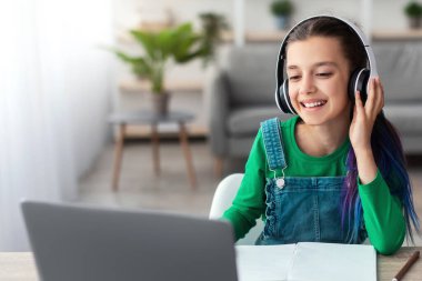 Modern Uzak Eğitim Konsepti. Genç kız portresi evde masada oturuyor, dizüstü bilgisayar kullanıyor, kablosuz kulaklık takıyor ve dokunuyor. Okullu kız salgın sırasında sanal ders alıyor.