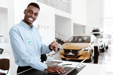 Gülümseyen siyah araba satıcısının portresi otomobil galerisinde dizüstü bilgisayar kullanırken el hareketi yapıyor. İş yerinde poz veren neşeli Afro yönetici, otomobil galerisindeki kameraya bakıyor.