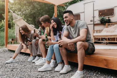 Mutlu çok uluslu arkadaşlar bira içiyor, sohbet ediyor ve gülüyor, karavanın yanında oturuyor, kamp yaparken eğleniyorlar. Yaz tatili kaçamakları konsepti