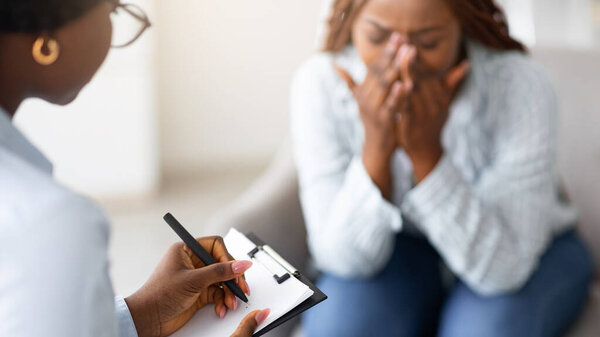 Stressed African American woman consulting psychologist, crying at medical office, selective focus. Psychiatrist helping desperate black lady with nervous breakdown, taking notes during session