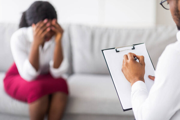 Unhappy young black lady crying on consultation with psychotherapist at clinic. Depressed Afro woman with mental problems expressing negative emotions at counselors office