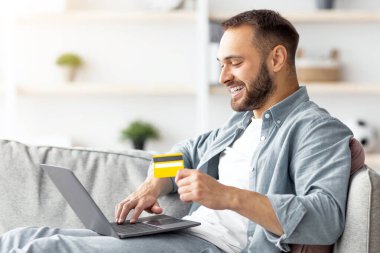 İnternetten alışveriş. Bilgisayarı ve kredi kartıyla evdeki koltukta oturan ve web mağazasından ürün alan beyaz bir gencin yan görüntüsü. Milenyum adamı internetten bir şeyler alıyor.