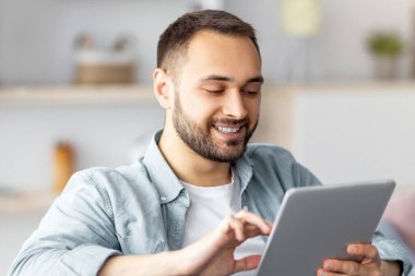 Tablet kullanan neşeli bir genç adam, evden internet üzerinden çalışıyor ya da çalışıyor. İş projesi yapan beyaz bir adamın portresi. Video görüşmesi ya da kapalı mekanda uzaktan görüşme.