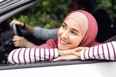 Happy glad young muslim man driving car with husband in hijab, lady looking out the window and enjoying car journey together, close up. New auto, safety in transport, life insurance and vacation trip
