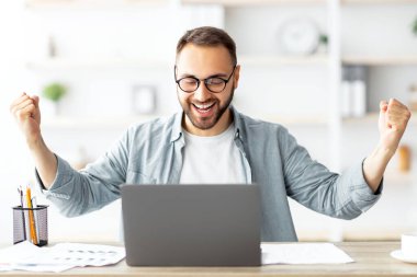 Genç beyaz adam laptopun önünde EVET işareti yapıyor, ev ofisinde başarıyı kutluyor. Milenyum adamı taşınabilir bilgisayarla çalışıyor, harika bir anlaşma yapıyor, online kontrat imzalıyor, başarı için heyecanlanıyor.