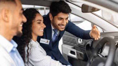 Mutlu ortadoğu milenyumu çifti galeride yepyeni bir arabanın içinde oturmuş, neşeli genç pazarlamacıları dinliyor, ön panele bakıyor ve gülümsüyor, yan manzara, yakın çekim, araba penceresinden çekiliyor.