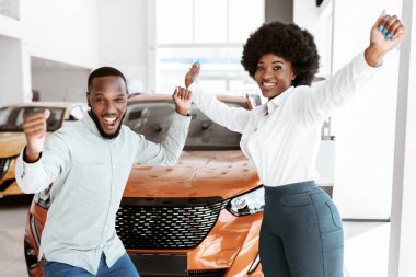 Heyecanlı Afro-Amerikan çift, başarılı araba alışverişini kutluyor, araba galerisinde dans ediyor ve eğleniyor. Coşkulu siyahi aile otomobil galerisinden yeni bir araç alıyor.