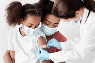 Afrika kökenli Amerikalı anne ve tatlı kız klinikte aşı yapıyorlar, koruyucu maskeler takıyorlar. Doktor, Coronavirus 'a iğne yaptıktan sonra omzuna bant takıyor.