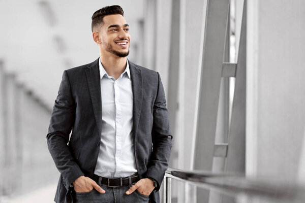 Handsome Young Middle Eastern Man Waiting Flight At Airport Terminal, Smiling Millennial Arab Businessman Standing With Hands In Pockets And Looking At Window, Ready For Trip, Copy Space