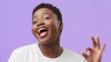 Tombul, genç bir Afrikalı Amerikalı kadın parlak mor bir arka plana karşı sevinçle poz veriyor. Elleriyle neşeli bir jest yapıyor ve neşeli bir ifade sergiliyor..