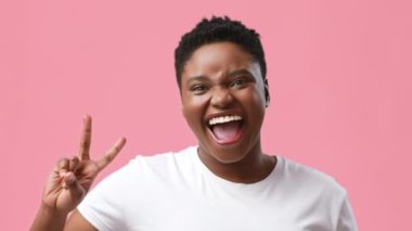Tombul vücutlu neşeli bir Afrikalı Amerikalı kadın geniş bir gülümsemeyle barış işareti veriyor. Canlı pembe bir geçmişi olan bir stüdyo ortamında pozitiflik ve güven yayıyor..