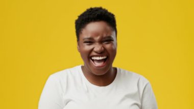 Neşeli bir ifadeyle genç bir Afrikalı Amerikalı kadın izole edilmiş bir stüdyoda parlak sarı bir arka plana gülüyor. Tombul yüz hatları neşeli tavrını geliştiriyor..
