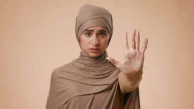 Başörtüsü takan genç bir Orta Doğulu kadın düz bir zemine karşı duruyor ve bir mesaj iletmek için elini kaldırıyor. İfadesinde odaklanmış ve ciddi görünüyor..