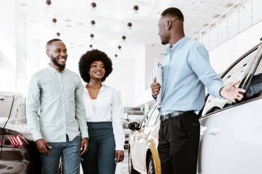 Şov salonundaki lüks araba hakkında neşeli bir Afro aile danışmanı. Mutlu genç siyah çift birlikte yeni bir araba satın alıyorlar, galerideki arkadaş canlısı satıcıyla konuşuyorlar.